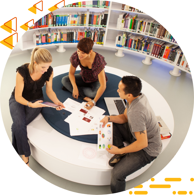 Aerial photo of three students collaborating on 设计 work while seated on a large ottoman, books are in the background.